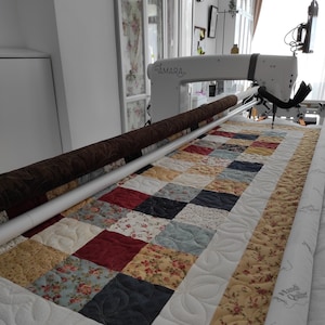 Puede incluir: Una máquina de acolchado de brazo largo blanca con una barra de acolchado marrón y una colcha con un diseño de patchwork de cuadrados en varios colores y patrones. La colcha se está acolchando con un diseño de hojas. La máquina tiene la palabra "AMARA" impresa en ella. La colcha tiene las palabras "Handi Quilter" impresas en ella.