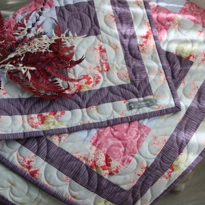 Può includere: Una coperta trapuntata con un design a patchwork che presenta motivi floreali in tonalità di rosa, bianco e verde. La coperta ha un bordo viola con un bordo smerlato e una piccola etichetta che recita "Kimber's Quilts".