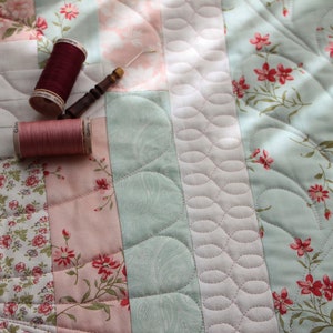 Könnte beinhalten: Nahaufnahme einer Steppdecke mit einem Blumenmuster in Rosa-, Grün- und Weißtönen. Zwei Garnrollen, eine rote und eine rosafarbene, liegen auf der Steppdecke.