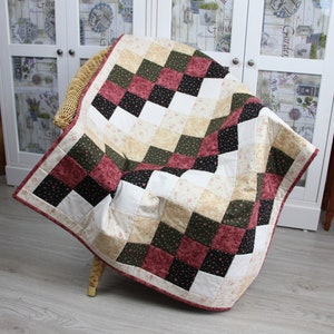 Può includere: Una trapunta patchwork con un motivo a scacchi in tonalità di rosso, verde, nero e bianco. La trapunta è realizzata con diversi tessuti con motivi floreali ed è piegata su una sedia in vimini.