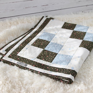 Peut inclure: Une courtepointe patchwork pliée avec un motif en damier dans des tons de vert, blanc et bleu. La courtepointe a un imprimé floral sur les carrés verts et une petite étiquette blanche avec le mot "Handmade" dessus.