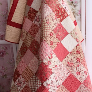 Könnte beinhalten: Ein Patchwork-Quilt mit einer Vielzahl von roten und cremefarbenen Blumen- und geometrischen Mustern. Der Quilt besteht aus Quadraten und Rauten mit einem cremefarbenen Rand. Der Quilt ist gefaltet und zeigt das gesteppte Design.