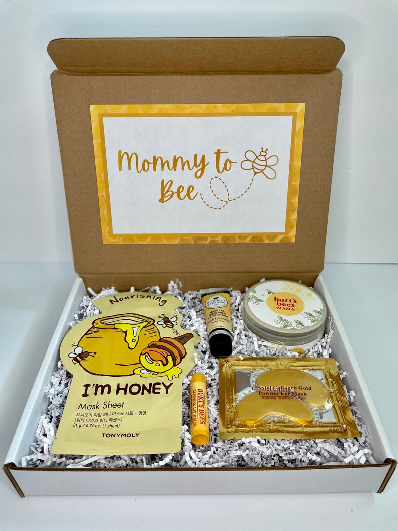 May include: A gift box labeled "Mommy to Bee" contains skincare products. Items include a honey-infused face mask, lip balm, eye masks, hand cream, and a round container of Burt's Bees Mama products, all nestled in white paper filler.