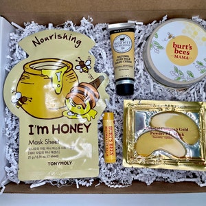 May include: A gift box filled with beauty products. Includes a honey-themed face mask, hand cream, lip balm, eye masks, and a jar of Burt's Bees Mama cream. The products are nestled in white paper filler.