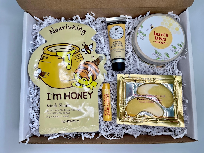 May include: A gift box filled with beauty products. Includes a honey-themed face mask, hand cream, lip balm, eye masks, and a jar of Burt's Bees Mama cream. The products are nestled in white paper filler.