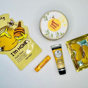 May include: A collection of skincare products, including a honey-themed face mask, a Burt's Bees Mama cream, a lip balm, a hand cream, and a gold eye mask. The products are arranged on a white surface.