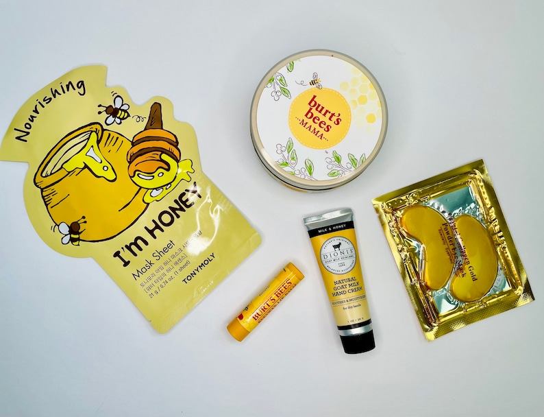 May include: A collection of skincare products, including a honey-themed face mask, a Burt's Bees Mama cream, a lip balm, a hand cream, and a gold eye mask. The products are arranged on a white surface.