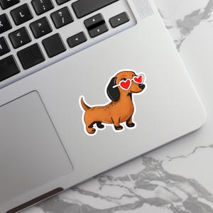 May include: A sticker featuring a cartoon dachshund wearing heart-shaped sunglasses. The dog is brown with black ears and paws. The sunglasses have red heart lenses and white frames. The sticker is placed on a laptop.