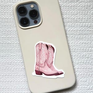 May include: A sticker of pink cowboy boots with white decorative stitching and small heart accents. The sticker is placed on a light beige phone case.