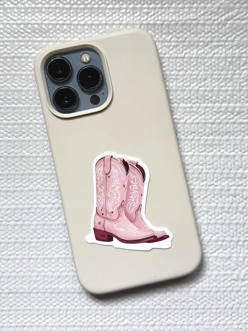 May include: A sticker of pink cowboy boots with white decorative stitching and small heart accents. The sticker is placed on a light beige phone case.