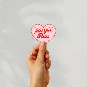 May include: A pink heart-shaped sticker with the text "Hot Girls Run" in red script. The sticker is held up by a hand with a gold ring. The sticker has a white border and small star accents.