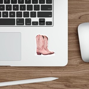 May include: A sticker featuring a pair of pink cowboy boots with intricate white floral and heart designs. The boots have a pointed toe and a low heel. The sticker is placed on a white surface next to a laptop keyboard.