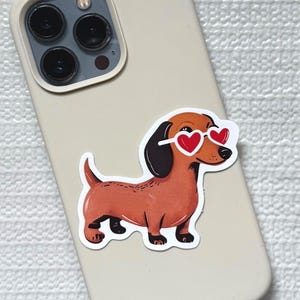 May include: A sticker of a brown dachshund wearing heart-shaped sunglasses is affixed to a beige phone case. The dog has black ears and paws. The phone case has a triple-lens camera.