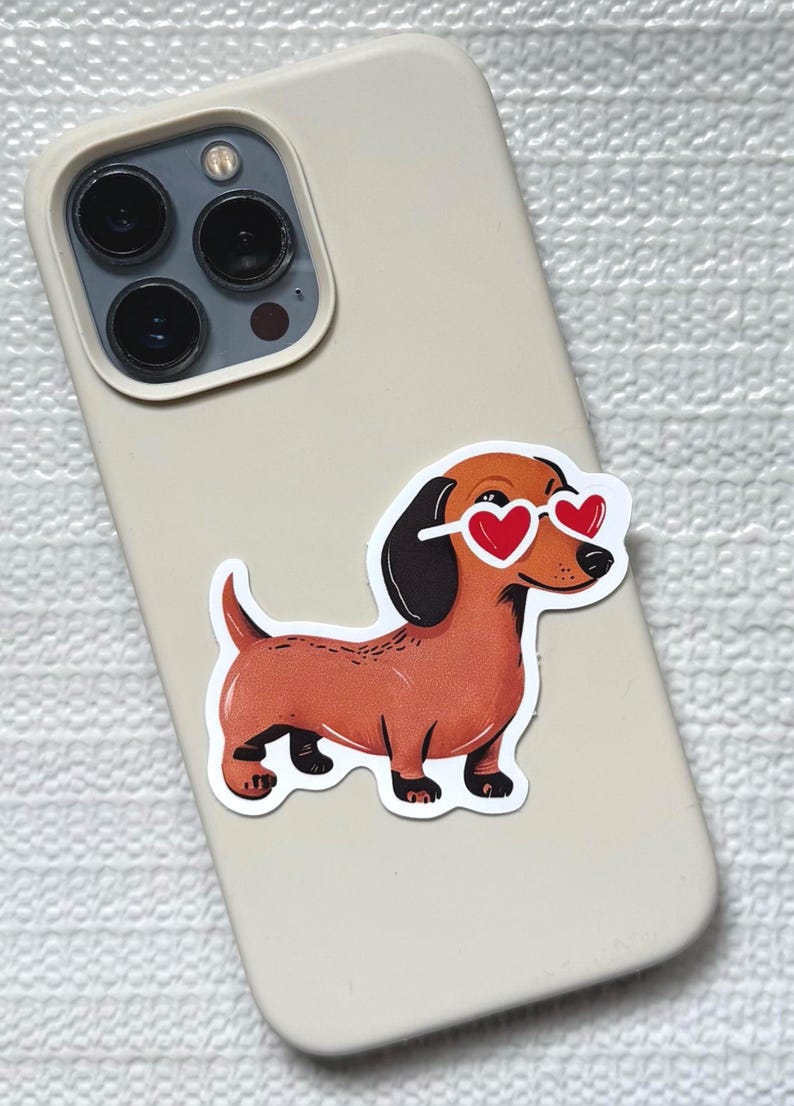 May include: A sticker of a brown dachshund wearing heart-shaped sunglasses is affixed to a beige phone case. The dog has black ears and paws. The phone case has a triple-lens camera.