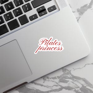 May include: A white sticker with the words "Pilates princess" in red script lettering. The sticker is on a silver laptop, with a marble-patterned surface visible in the background.