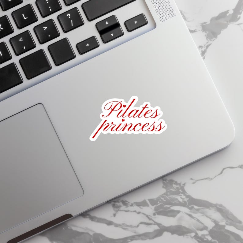 May include: A white sticker with the words "Pilates princess" in red script lettering. The sticker is on a silver laptop, with a marble-patterned surface visible in the background.