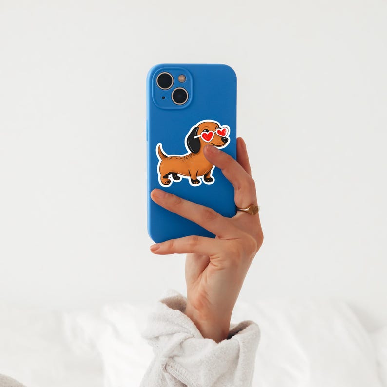 May include: A blue smartphone case with a sticker of a cartoon dachshund wearing heart-shaped sunglasses. The dachshund is brown with a black ear and tail. The phone case is held in a hand with a gold ring.