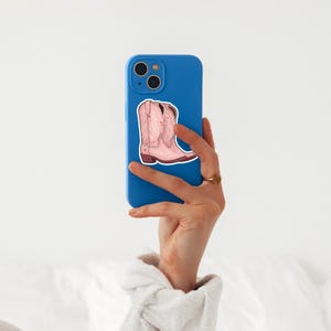 May include: A sticker of a pink cowboy boot with decorative stitching is affixed to a blue phone case. The boot has a brown sole and heel. The phone is held up against a white background.