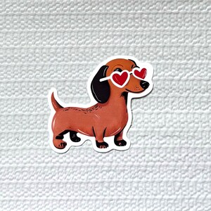May include: A sticker featuring a cartoon dachshund wearing heart-shaped sunglasses. The dog is brown with black ears and paws. The sunglasses are white-framed with red heart lenses. The sticker is on a white textured background.