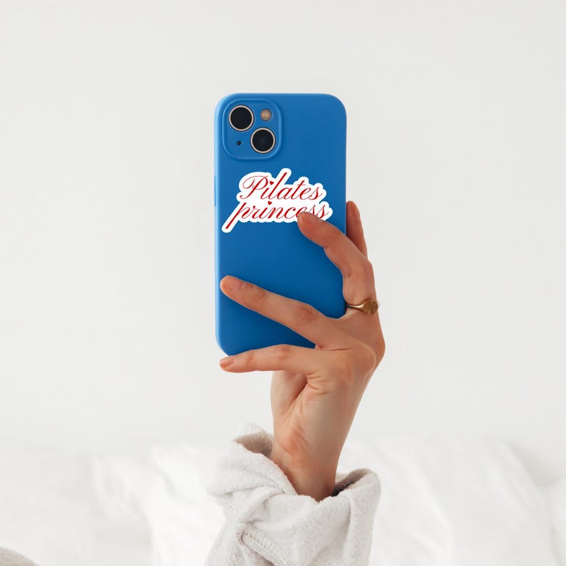 May include: A blue smartphone with a white and red "Pilates princess" sticker. The phone is held in a hand with a gold ring. The background is a soft white.