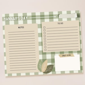 May include: A planner with a green and white gingham pattern, with sections for notes, to-do lists, and daily goals. The text "HANNAH'S DAY" is at the top, with decorative leaves at the bottom. A pen, phone, and notebook are in the background.