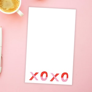 May include: A white notepad with the word "XOXO" printed in pink and red letters at the bottom. The notepad is on a pink surface with a cup of coffee and pens.