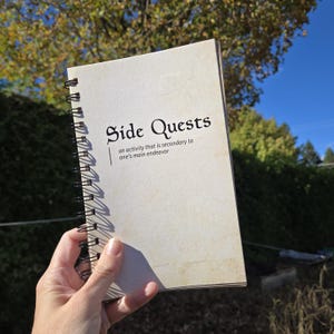 May include: A spiral-bound notebook with a cream-colored cover featuring the black text Side Quests and the definition an activity that is secondary to one's main endeavor printed below it. The notebook has a black metal wire binding on the left side.