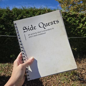 May include: A spiral-bound notebook with a cream-colored cover featuring the black text Side Quests and the definition an activity that is secondary to one's main endeavor. The notebook has a black metal coil binding along the left side.