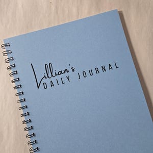 May include: A light blue spiral-bound journal with the text "Lillian's DAILY JOURNAL" in black. The journal is open, displaying the cover. The spiral binding is black.