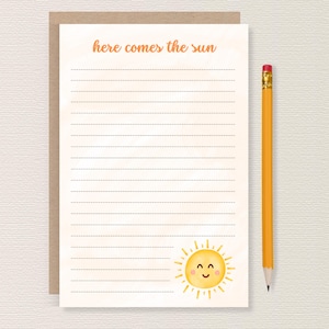 May include: A notepad with the text "here comes the sun" in orange script. The notepad has lined pages and a smiling sun illustration. A yellow pencil with a pink eraser rests beside the notepad.