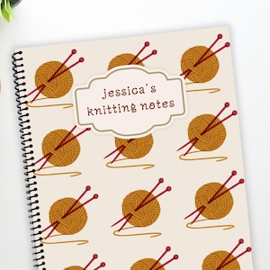 May include: A cream-coloured spiral-bound notebook with a pattern of yarn balls and knitting needles. The notebook has the text "Jessica's knitting notes" in a decorative frame. The design is in shades of brown and red.