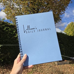 May include: A light blue spiral-bound journal with black text on the front cover that reads Lillian's DAILY JOURNAL. The notebook features a solid matte blue cover and a black metal wire binding along the left side for page turning.