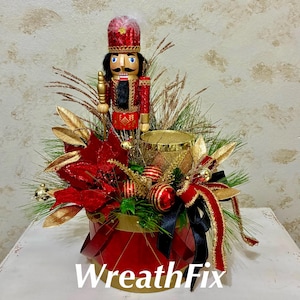 May include: A decorative arrangement featuring a red nutcracker figure, a gold drum, red poinsettia flowers, and pine needles. The base is a red drum-shaped container with gold trim, accented by red and black ribbons and gold-toned ornaments.