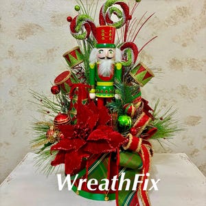 May include: A Christmas centerpiece featuring a central nutcracker figure in a green and red uniform, surrounded by red poinsettia flowers, green pine needles, red and green ornaments, and spiral candy decorations, with the text WreathFix.