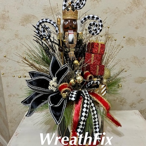 May include: A decorative arrangement featuring a nutcracker figure with a gold crown, black and white checkered accents, red glittered gift boxes, a large black flower with white trim, gold sprigs, and ribbons, all set in a container labeled WreathFix.