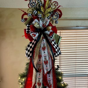 May include: A Christmas tree topper featuring a large bow with long ribbons in red, black, white, and gold. The ribbons display a nutcracker pattern and a black and white checkered design. The topper includes red and gold decorative sprays.