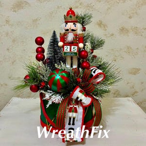 May include: A Christmas-themed decorative arrangement featuring a nutcracker figure with a countdown calendar block displaying 25, surrounded by green pine branches, red ornaments, a striped ball, and a ribbon with a nutcracker print.
