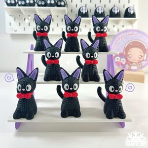 Puede incluir: Una colecci&oacute;n de figuras de gatos negros con orejas moradas y pajaritas rojas, dispuestas en una exhibici&oacute;n escalonada blanca y morada. Cada gato tiene grandes ojos blancos con una pupila negra. Figuras de gatos adicionales son visibles en el fondo.