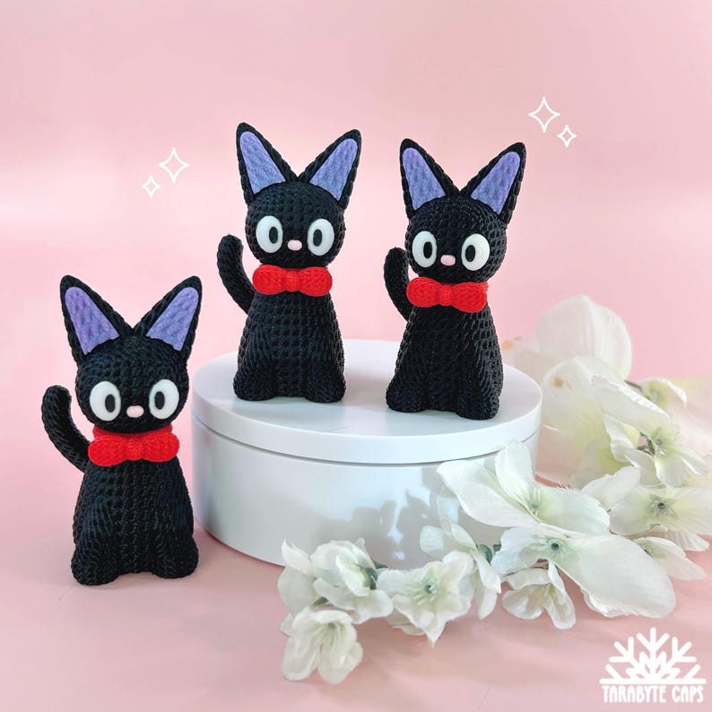 Puede incluir: Tres figuras de gatos negros con pajaritas rojas y ojos blancos grandes se exhiben sobre una plataforma blanca. Los gatos tienen orejas forradas de morado y posan con una pata levantada. El fondo es rosa suave con flores blancas.