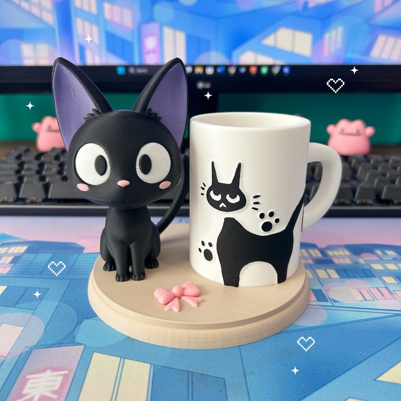 May include: A black cat figurine with purple ears and a white-eyed expression sits beside a white mug with a black cat design. A small pink bow and a wooden base complete the set. The mug features a black cat silhouette.