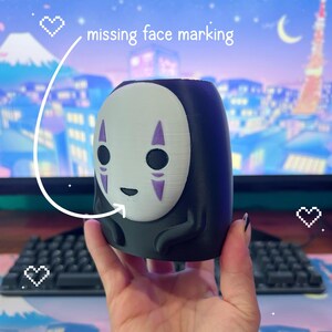 Puede incluir: Un macetero negro y blanco del personaje Sin Rostro con detalles morados, sostenido en una mano. El macetero tiene forma redondeada y presenta el dise&ntilde;o distintivo de la m&aacute;scara del personaje. El texto "missing face marking" es visible.