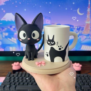 May include: A black cat figurine with large white eyes and purple ears sits on a wooden base next to a white mug. The mug features a black cat design. A pink bow and two pink figures are also on the base.