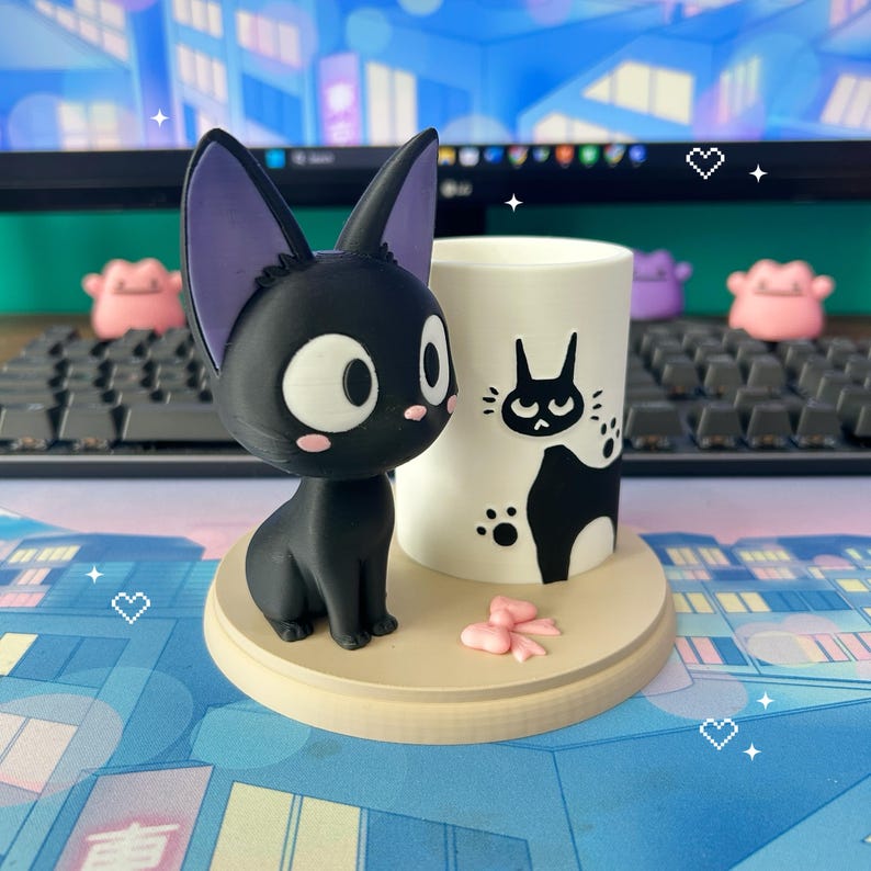May include: A black cat figurine with purple inner ears and large white eyes sits beside a white mug with a black cat graphic. The mug and cat are on a round, light brown base. Pink decorative accents are also present.