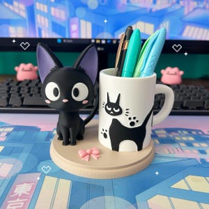 May include: A black cat figurine with purple ears sits beside a white mug decorated with a black cat silhouette. The mug holds pens and markers. A small pink bow and a round wooden base complete the desk organiser.