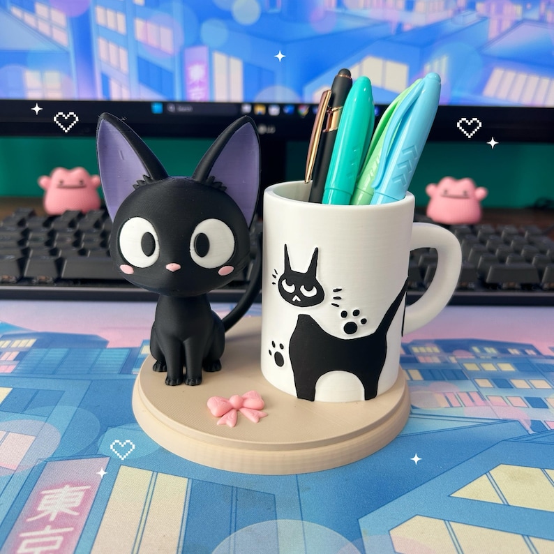 May include: A black cat figurine with purple ears sits beside a white mug decorated with a black cat silhouette. The mug holds pens and markers. A small pink bow and a round wooden base complete the desk organiser.