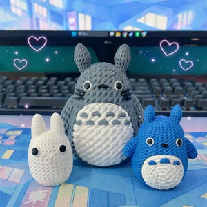 May include: A collection of handmade crochet figures, including a large gray Totoro, a small blue Totoro, and a white bunny-like creature. The figures have black eyes and are displayed on a blue surface with a keyboard in the background. Decorative hearts are also present.