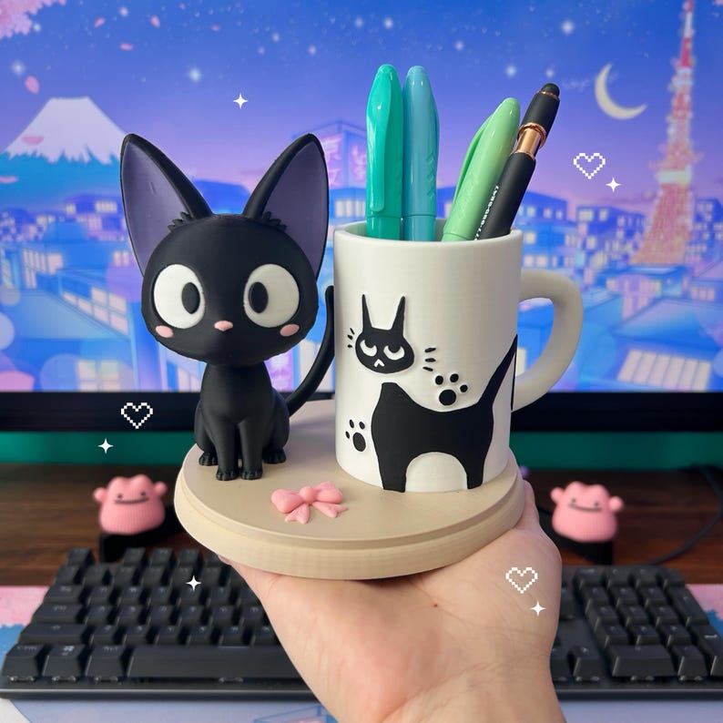 May include: A black cat figurine sits beside a white mug featuring a black cat design. The mug contains green and blue pens. The cat and mug are on a wooden base with a pink bow. A keyboard is in the foreground.