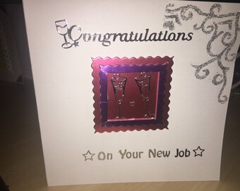 May include: A white card with a silver glitter design in the top right corner. The card says "Congratulations" in silver glitter lettering with a silver glitter design of two wine glasses. The card also has a red and purple square with a silver glitter design of two wine glasses with hearts. The card says "On Your New Job" in silver glitter lettering with a silver glitter star on each side.
