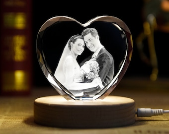 Puede incluir: Un adorno de cristal en forma de corazón con una foto en blanco y negro de una pareja en su interior. El adorno está sobre una base de madera con una luz debajo.
