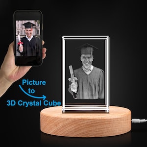 May include: A 3D crystal cube with a black and white photo of a person in a graduation cap and gown holding a diploma. The cube is on a wooden base with a light cord.
